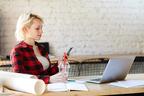 Side view portrait of blonde young woman wearing casual shirt using smartphone while working in modern office, copy space