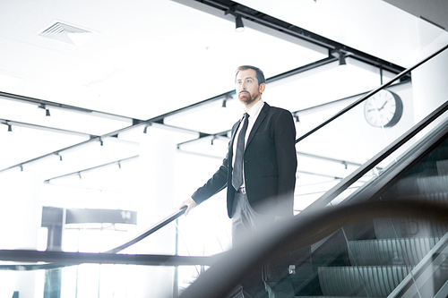 Serious businessman in elegant suit moving downwards on escalator to leave airport