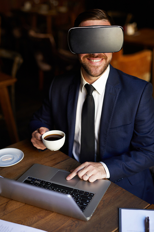 Happy successful young bearded businessman in virtual reality goggles using laptop and drinking black coffee while working in restaurant