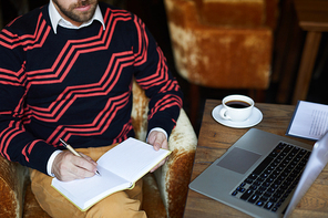 Young contemporary author sitting in armchair in cafe and writing down his ideas in notebook by cup of coffee