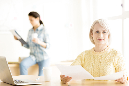 Young pretty female sitting with cup laptop and papers  with brunette working on back in soft focus