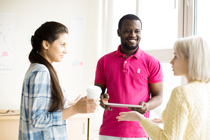 Two young women and black man maintaining conversation on coffee break standing in office