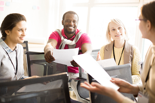 Two young women looking at female speaker and smiling black man holding out paper  sitting on chairs in office