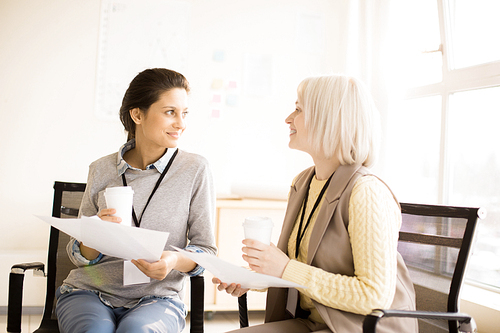 Side view of beautiful smiling ladies with badge on neck holding papers and take-out cups in hands looking at each other sitting on chairs