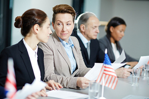 Mature delegate showing papers to political partner sitting next to her on background of other speakers