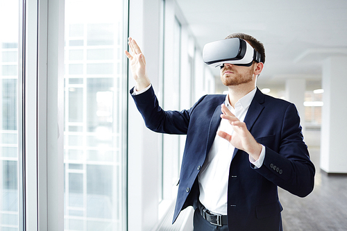 Contemporary young businessman in vr headset taking part in virtual conference while standing by window of office-center