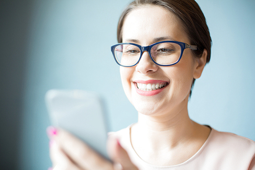Pretty businesswoman in glasses smiling and using smartphone while sitting near light blue wall.