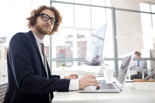 Young confident businessman in suit and eyeglasses  while sitting by desk and typing