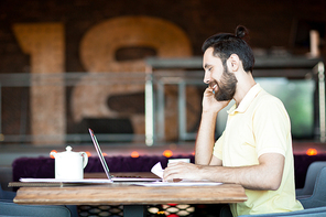 Happy young businessman sitting by table in cafe, talking by smartphone and browsing in the net