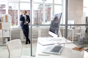 Young elegant businessman in suit standing by window in office and speaking by smartphone