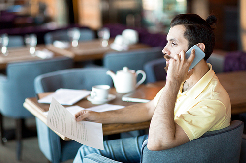 Contemporary employee with smartphone and paper sitting by table in cafe and phoning client