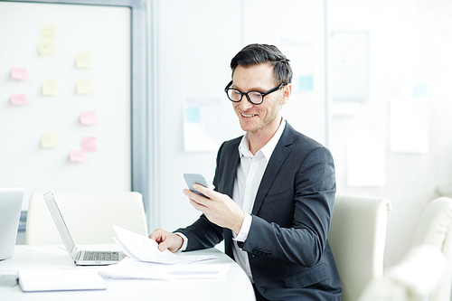 Businessman in eyeglasses text messaging on his cell phone while working with documents at office desk