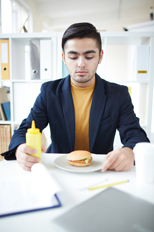 Young contemporary manager sitting by workplace and looking at hamburger on plate before having lunch