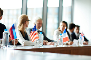 Focus on small national flags, bottles of water and glasses on conference table, politicians talking in background