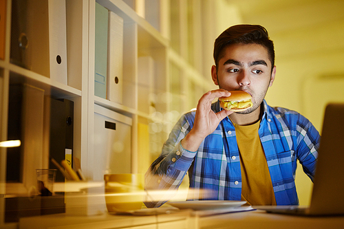 Hungry manager with hamburger in mouth watching webcast in laptop while staying in office for night