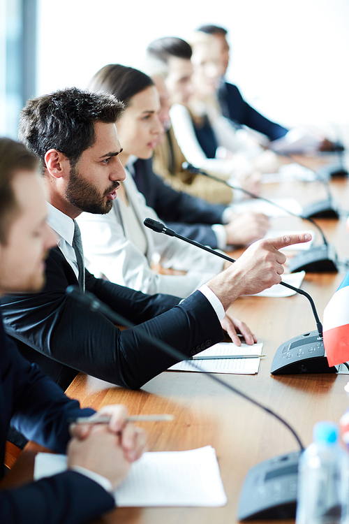 Aggressive confident handsome young bearded political leader in suit expressing his position and pointing with forefinger while speaking into microphone at international meeting