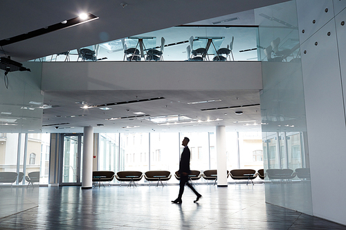 Profile view of confident businessman wearing suit walking along spacious office lobby with panoramic windows, motion shot