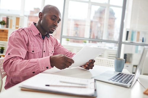 Young concentrated African American man sitting at office desk with laptop and reading important paper