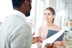 Pretty young manager and her bearded African American colleague standing at panoramic window of modern open plan office and analyzing statistic data, lens flare