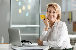 Pretty middle aged businesswoman sitting by workplace and holding sticker with drawn heart on valentine day