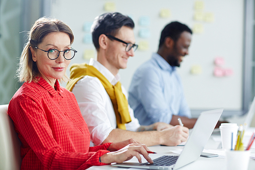 Confident mid-aged businesswoman in eyeglasses and red blouse on background of her team working