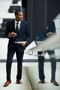 Young man in elegant suit standing on staircase and looking through window while messaging