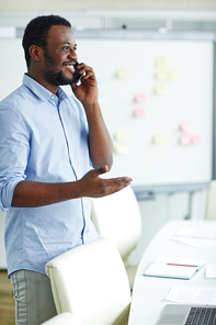 Happy young agent in shirt and jeans having phone talk with one of clients while working in office