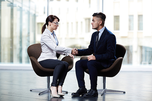 Confident entrepreneur with long dark hair shaking hand of her business partner while they sitting opposite each other, interior of modern boardroom with panoramic windows on background
