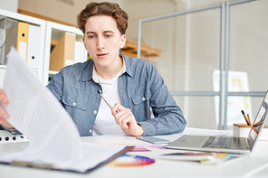 Young casual businessman sitting at his workplace and examining documents