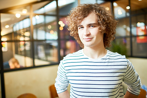 Young employee with curly hair looking at you with smile while working in his office