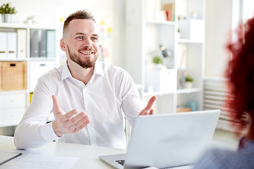 Young handsome bearded office manager in white shirt sitting at table with laptop, chatting with colleague and smiling