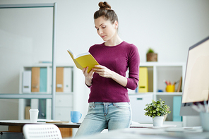 Young businesswoman standing near her workplace and reading a book at office