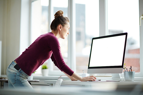 Young programmer or web-designer in purple pullover and blue jeans leaning over desk while reading online information on monitor display