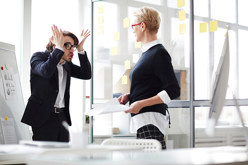 Emotional young manager standing at marker board and gesticulating vigorously while trying to explain something to his middle-aged superior, interior of open plan office on background