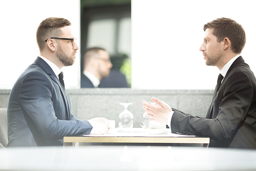 Side view of two young businessmen sitting by table in front of one another and discussing new business