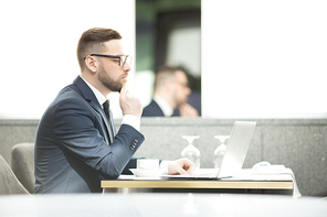 Pensive businessman sitting by table in front of laptop and analyzing online data