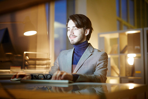 Smiling handsome designer sitting in front of computer and analyzing results of accomplished project, interior of dim office with panoramic windows on background