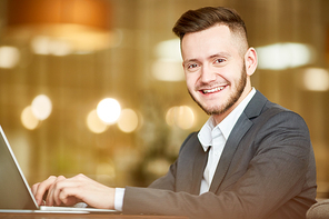 Portrait of young handsome Caucasian man in business suit sitting at laptop and smiling at camera cheerfully on blurred background