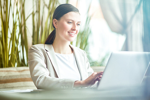 Young attractive Caucasian woman talking to business partner on laptop and smiling cheerfully while having lunch break in cafe