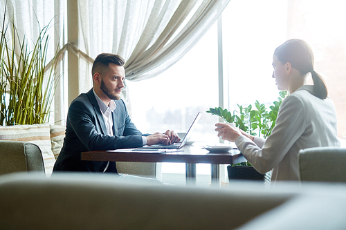 Young Caucasian bearded businessman sitting at cafe table with laptop and talking to his female coworker during lunch break