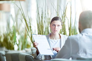 Young confident businesswoman showing clipboard with important paper to her male colleague at business lunch in cafe