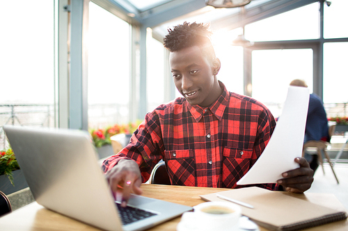 African-american guy in shirt sitting in cafe in front of laptop and preparing report for conference or watching webinar