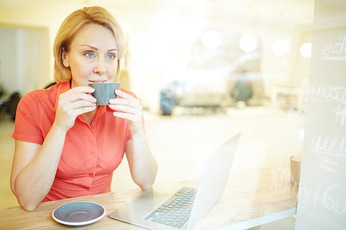 Mature blonde drinking hot fresh coffee from grey mug in cafe and looking through window