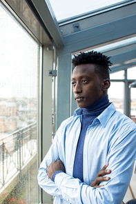 Young African-american man with crossed arms looking through window while it is raining outside