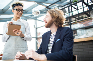 Happy young employee ordering his favorite drink or meal in cafe while waitress making notes