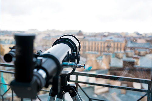 Professional telescope of black color by window with urban scene behind it