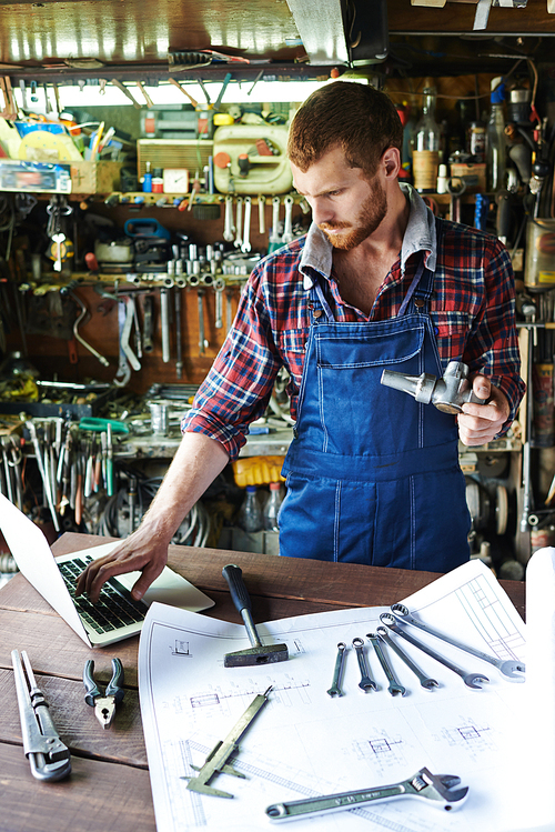 Portrait of handsome bearded mechanic wearing uniform overalls busy working in garage, assembling parts using tools and blueprints while looking down at laptop checking manual