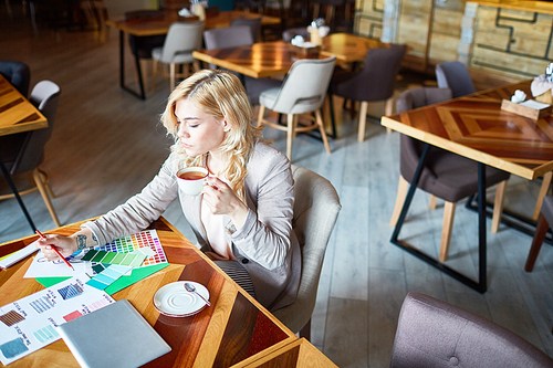 High angle view of confident blond-haired interior designer using color swatches while working on promising project, interior of spacious coffeehouse on background