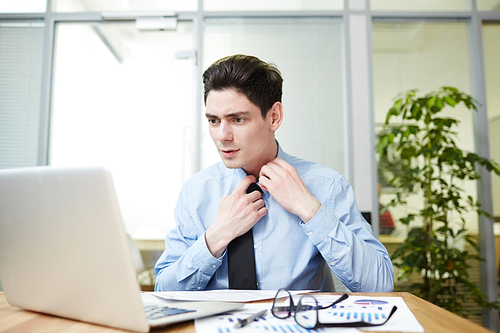 Exhausted young businessman loosening tie while sitting in front of laptop and analyzing statistic data, interior of spacious office on background