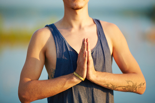 Close-up view of young man in tank top with tattoos meditating outdoors and holding hands in namaste